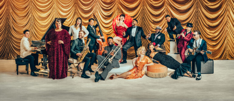 Photo os the members of post modern jukebox posing infront of a gold curtain with instruments