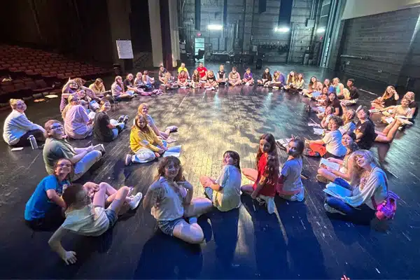 students sitting in a circle on stage during a summer camp